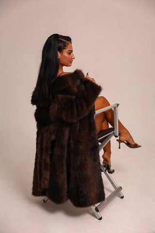 Woman wearing a long brown fur coat sitting on a chair against a plain background